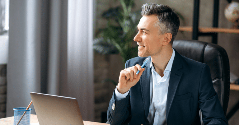 Smiling man thinking in a suit holds a pen at a desk with a laptop, looking right.